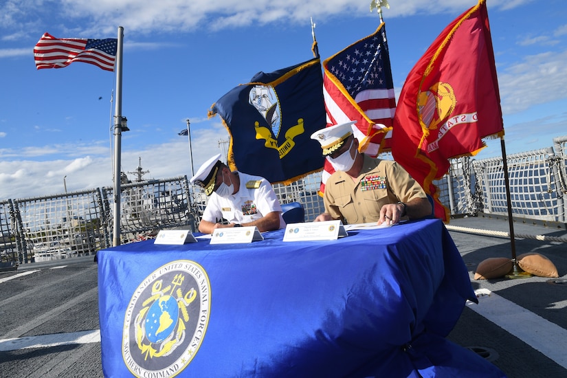 Two service members sit at a table; flags fly behind.