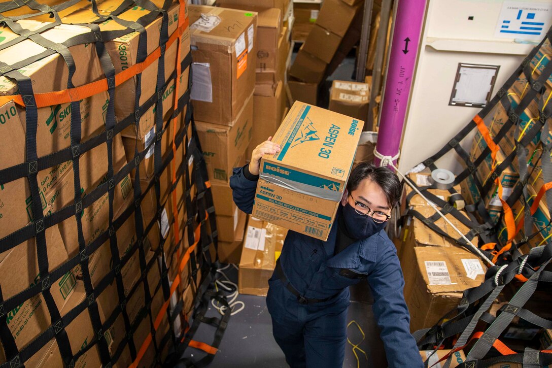 A sailor carries a box over his shoulder.