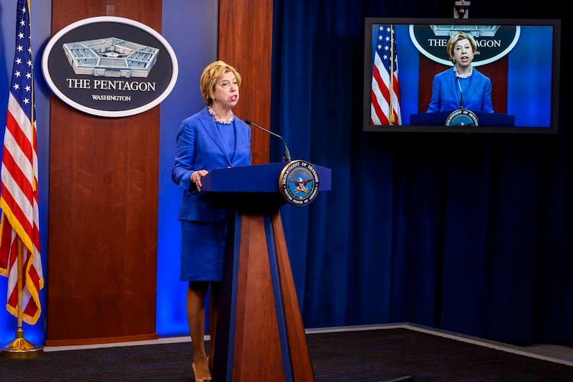 A woman in a blue suit stands at a lectern and speaks to a virtual audience. A sign behind her indicates that she is speaking from the Pentagon.