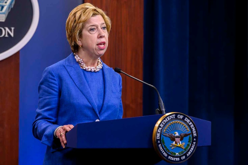 A woman in a blue suit stands at a lectern and speaks to a virtual audience.