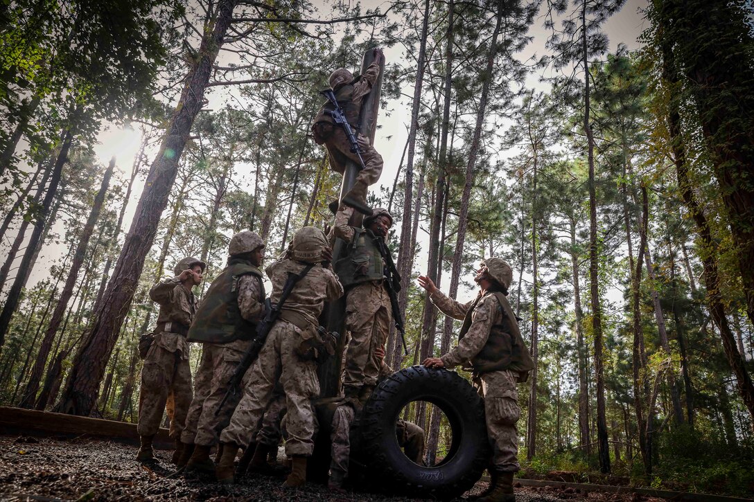 A group of Marines with weapons stand on each other to climb a pole.
