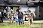 Soldiers walk with civilians away from an open helicopter in a field.