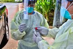 A person wearing personal protective equipment puts a swab into a vial being held by someone who is also wearing protective gear.