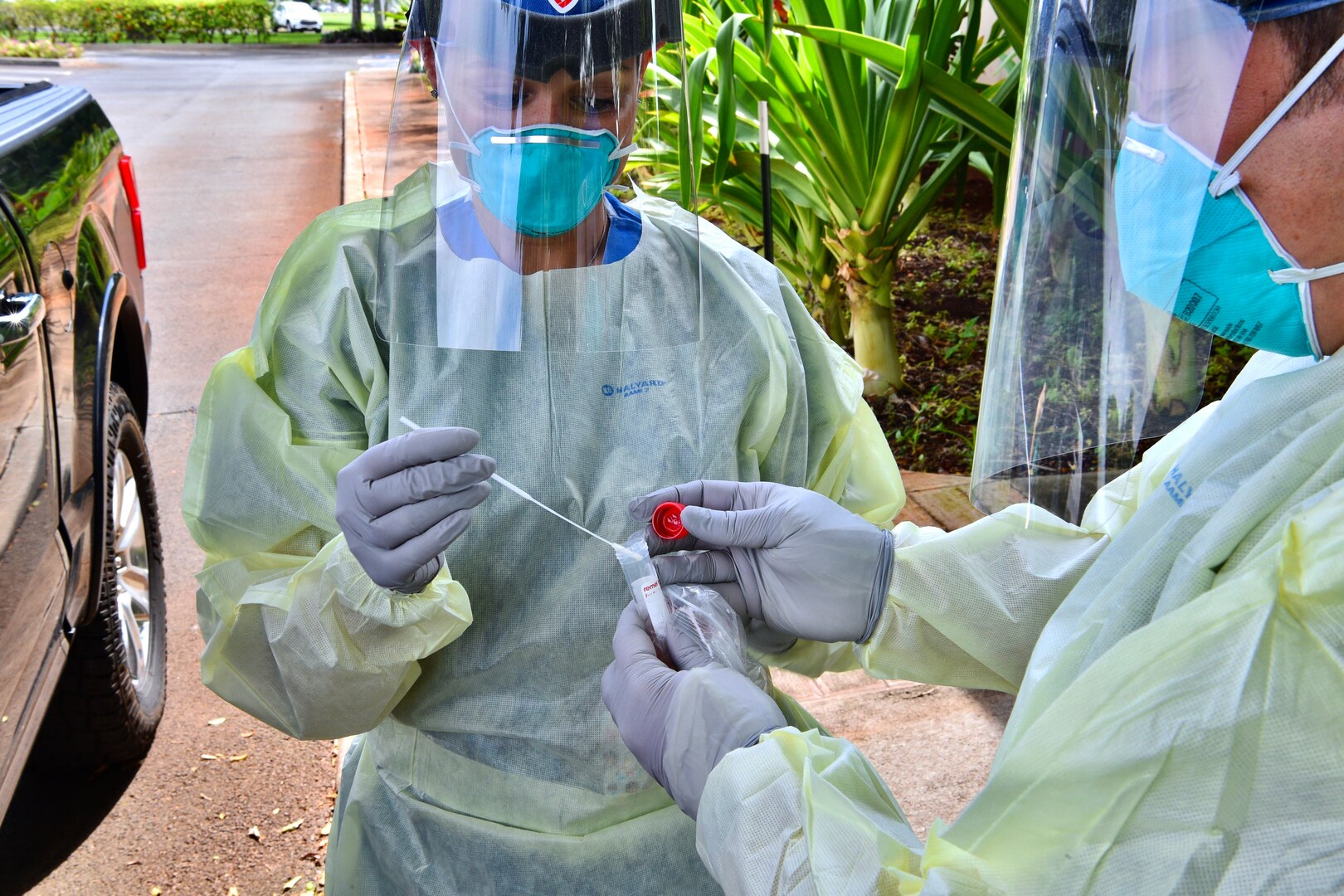 A person wearing personal protective equipment puts a swab into a vial being held by someone who is also wearing protective gear.