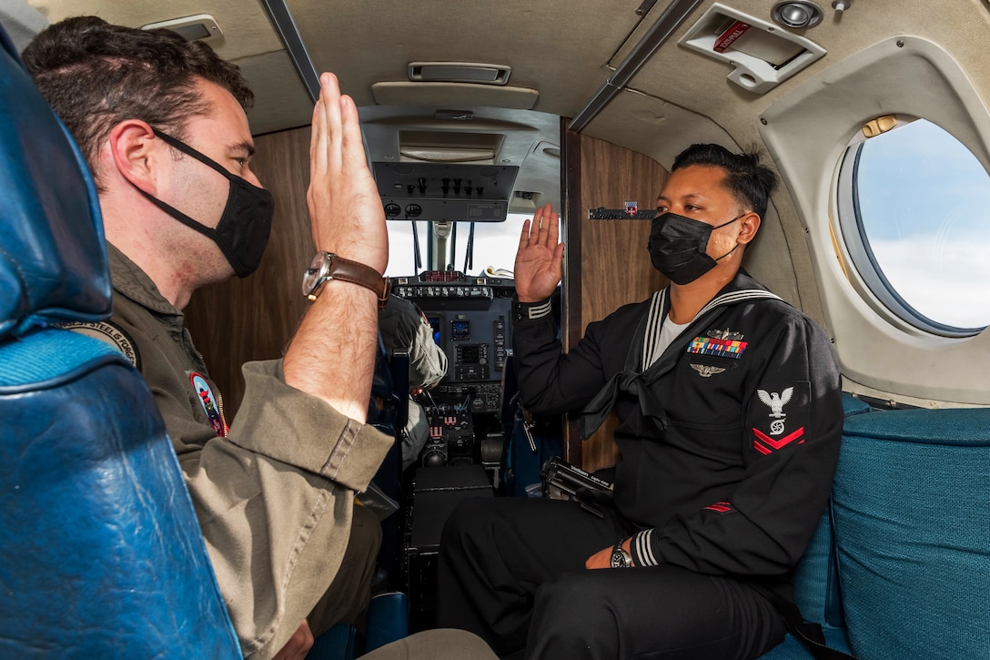 A Navy officer wearing a face mask administers the oath of enlistment to a sailor wearing a face while in-flight.