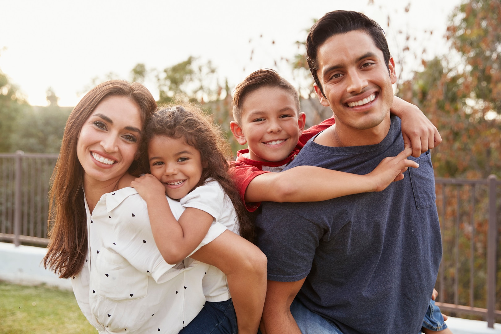 Young parents piggyback their children in the park, smiling to camera, focus on foreground