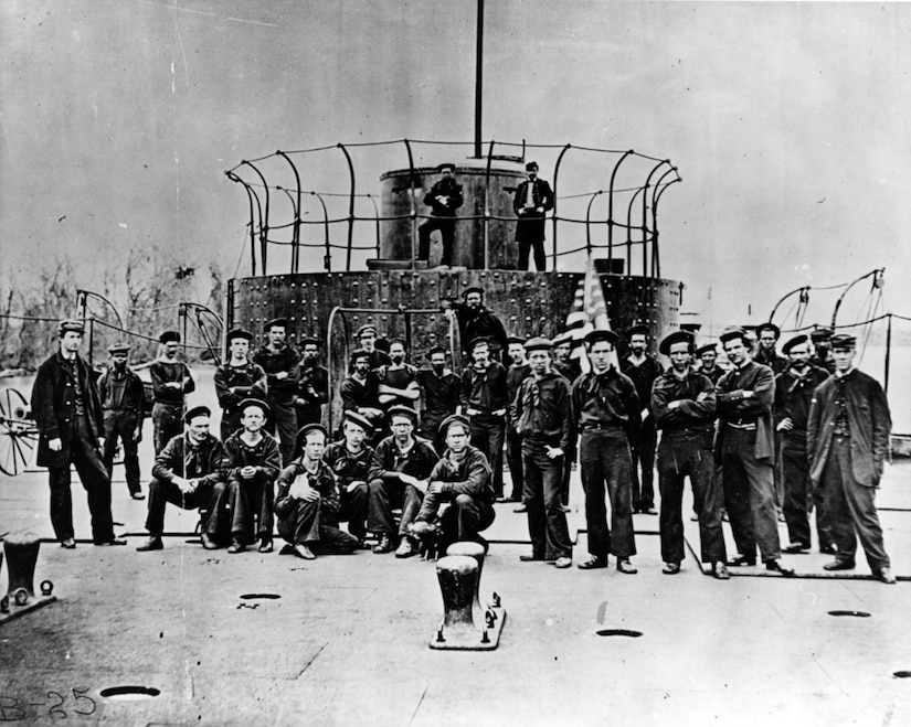An old photo shows dozens of sailors posing near the turret of a ship.