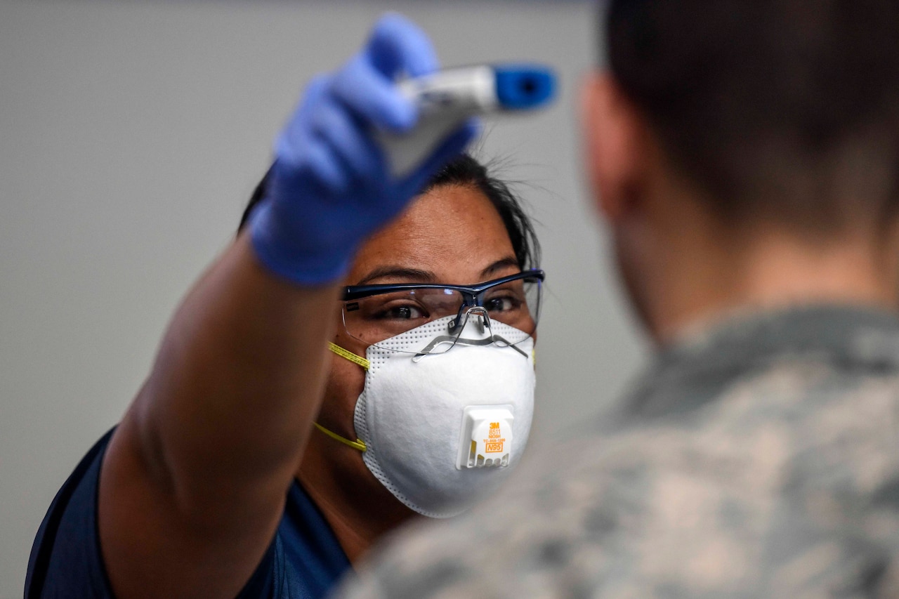 An airman wearing a face mask holds a thermometer to another airman's head.