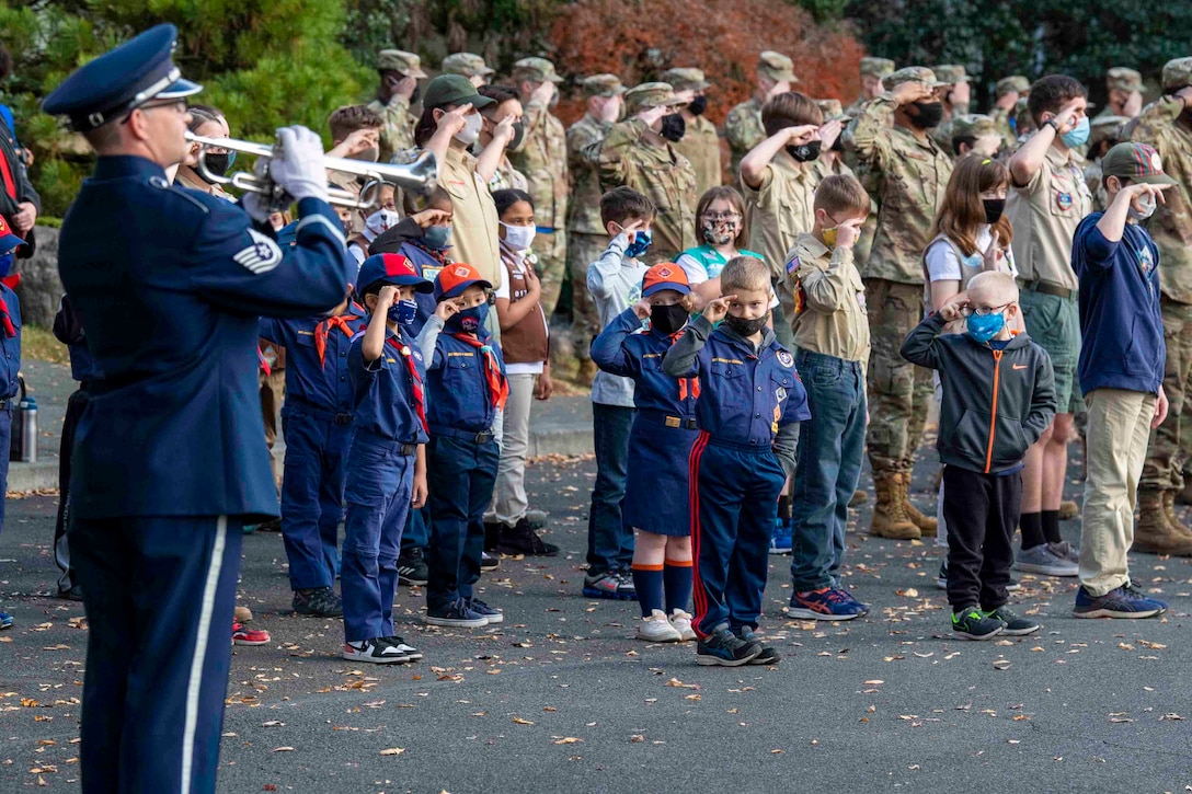 A group of young children salute an airman who is playing an instrument; a group of service members stand behind saluting.