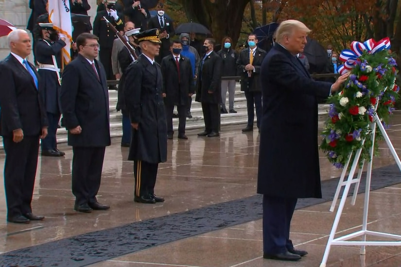 A man touches a wreath. In the background are multiple men and women;  some people wear military uniforms, and others are dressed in suits.