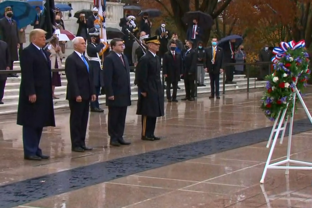 Four men stand in a line and face a wreath. In the background are multiple men and women; some wear military uniforms, and others are dressed in suits.