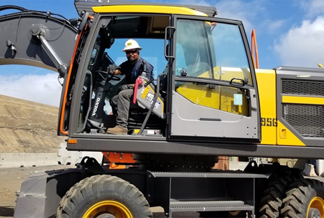 A man with a hard hat sit in the cab of a yellow piece of material handling equipment.