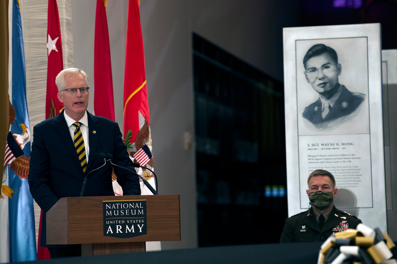 A man speaks from a podium, next to him sits a man in military uniform in front of a historical display.