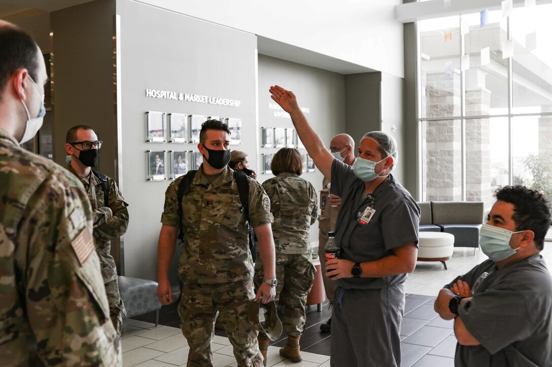 Two people wearing face masks and dressed in hospital uniforms talk to a group of people dressed in military uniforms, who are also wearing face masks.