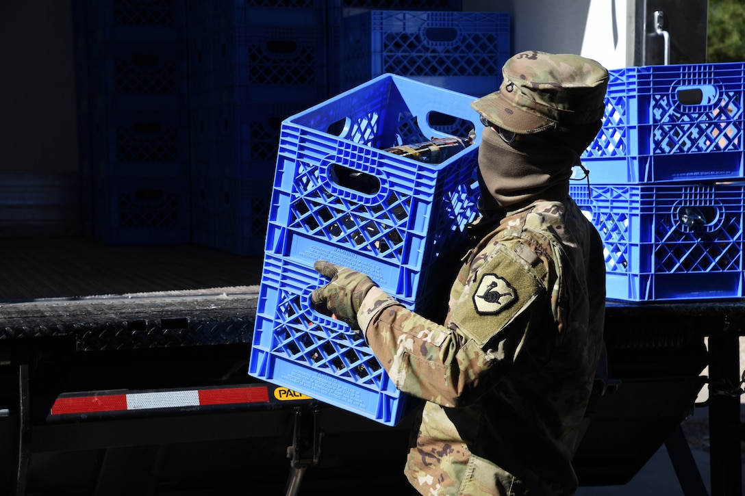 A national guardsman wearing a face mask and gloves puts two crates into the back of a large truck.