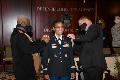 Two men change the rank on female uniform. One her husband, one her director