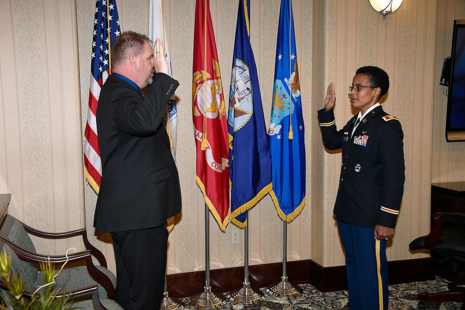 Man in suit and woman in uniform both stand in facing eachother in front of flags with their right hand raised