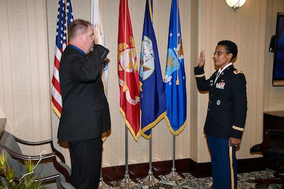 Man in suit and woman in uniform both stand in facing eachother in front of flags with their right hand raised