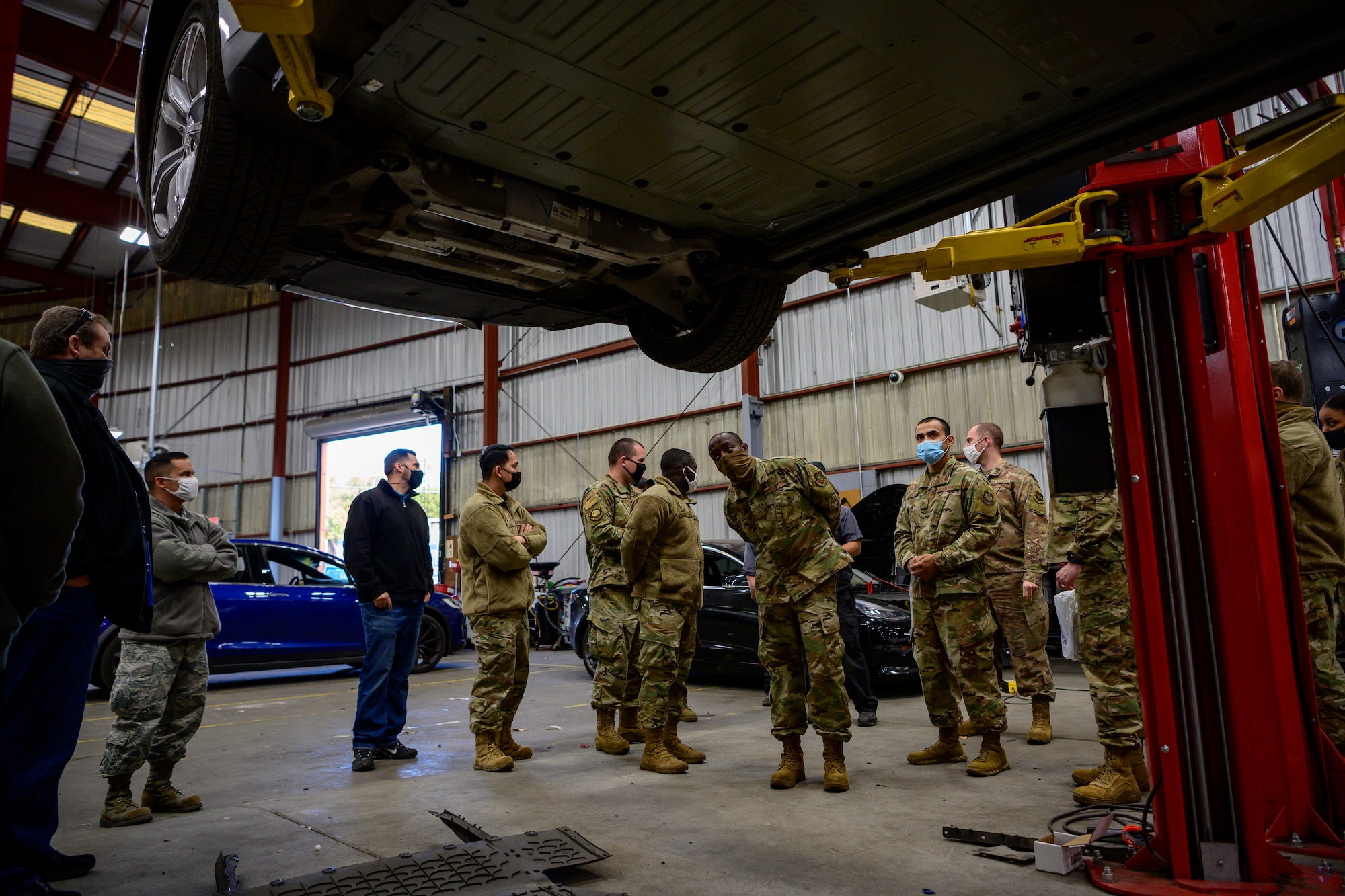 Airmen stand around a lifted Tesla vehicle inside a large warehouse.