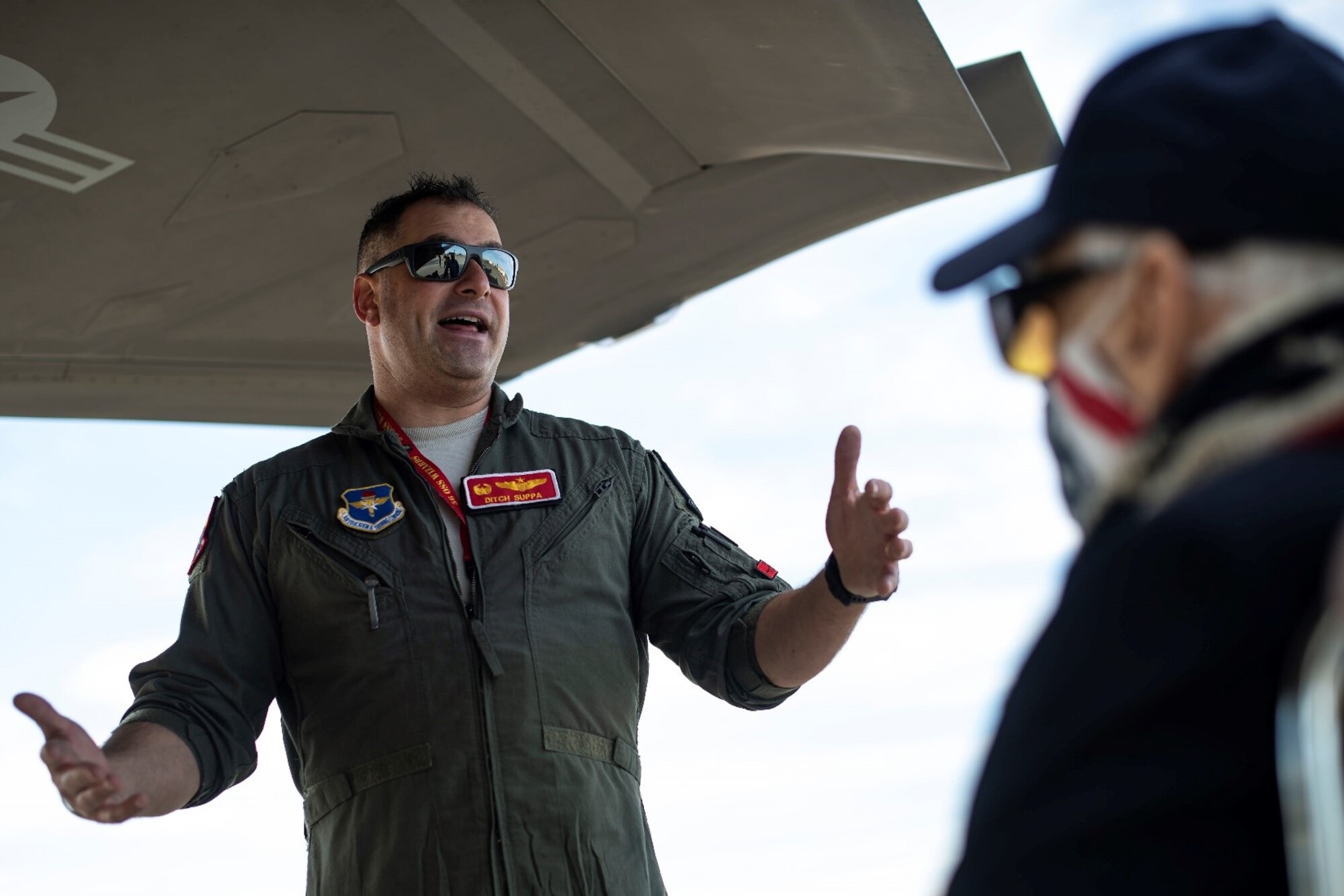 A military member in a flight suit speaks to Dean "Diz" Laird next to a fighter jet.