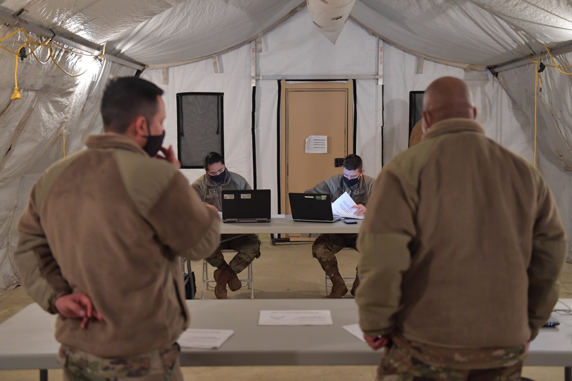 Two Airmen standing behind a table, while two other Airmen sit behind another table.