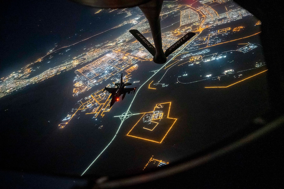 Two aircraft fly over a city illuminated by blue, white and orange lights.