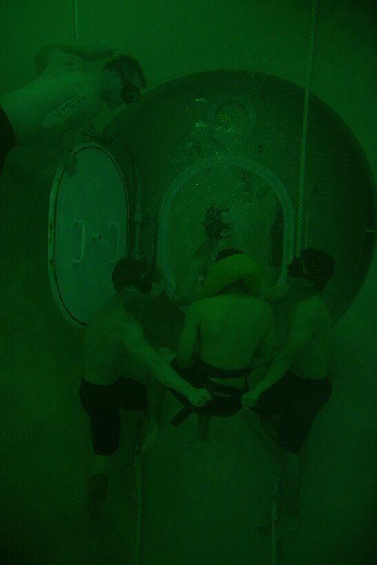 Two men hold onto straps attached to a third man facing an underwater portal in a large tank. Two other men watch.
