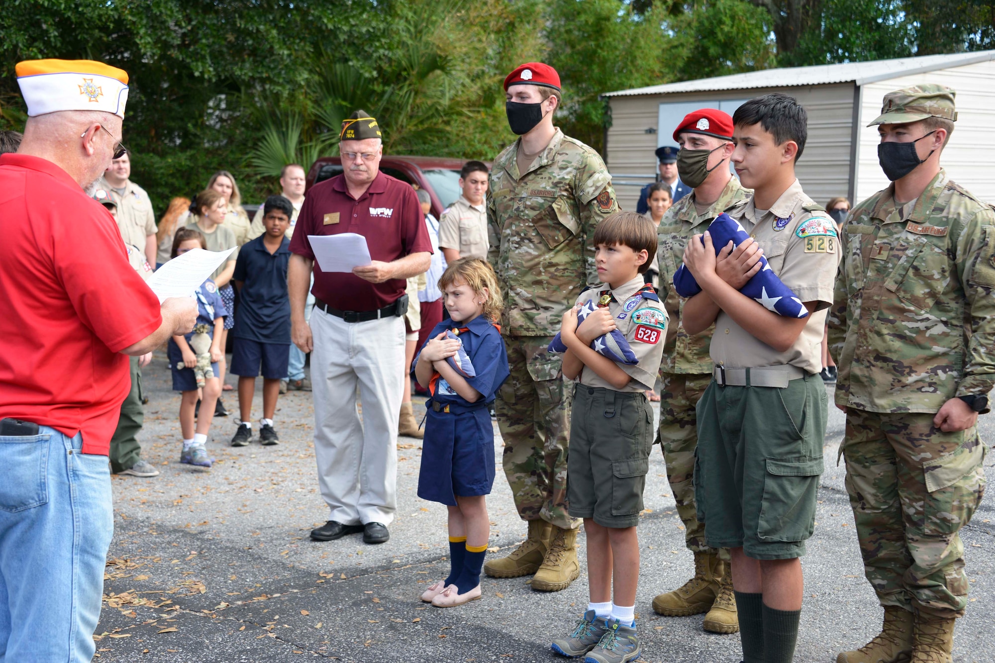 We stand off to the side as three children in boy and girl scout uniforms face a man in a red shirt and blue pants reading from a paper during a Veterans Day ceremony. Behind the two boys and a girl stand three Special Tactics Airmen at attention.To the children's right stands a man in a burgundy shirt and white pants reading along from a piece of paper.  In the distance we can see families standing and watching the ceremony.