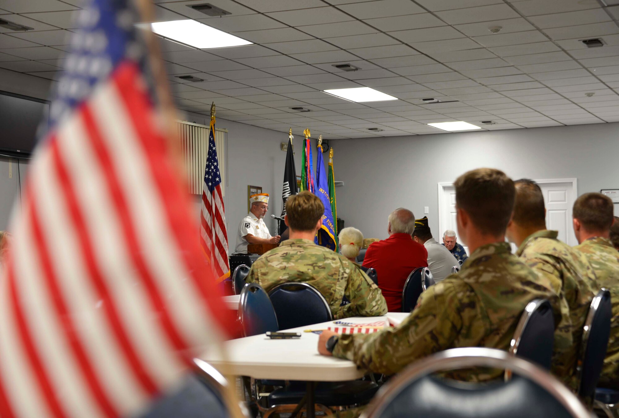 We look across a table at a room full of Veterans of Foreign War and Special Tactics Airmen seated at white tables. A Man in a white shirt stands at a podium across the room addressing those assembled for the Veterans Day ceremony.