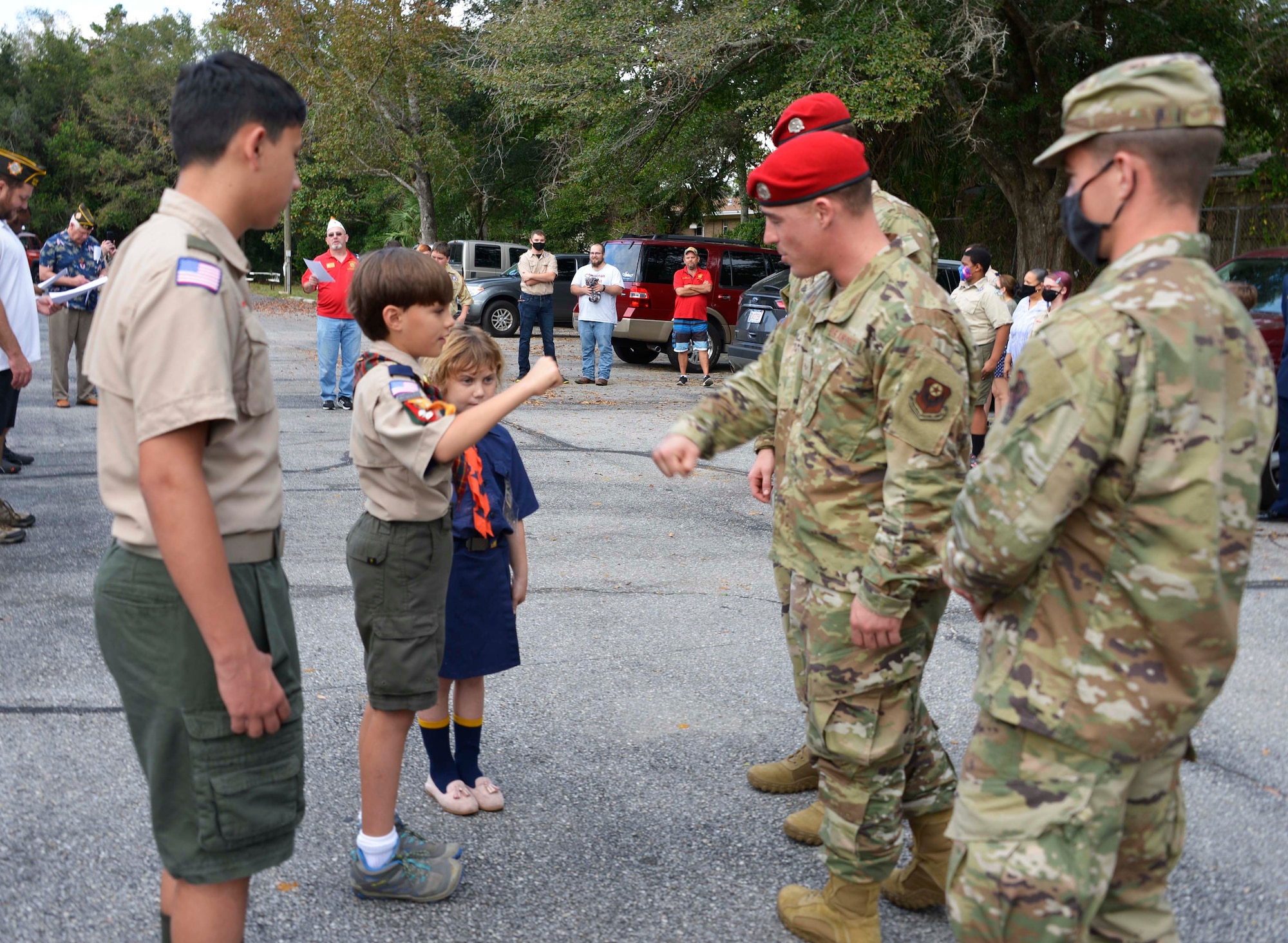 We look from the side as three children in boy and girl scout uniforms stand across from three Special Tactics Airmen. The middle of the three in each line are extending right elbows to greet each other in a COVID-19-approved greeting fashion during a Veterans Day ceremony.