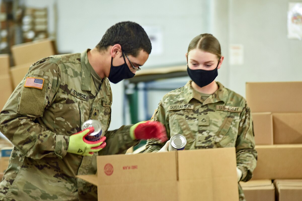 Two National Guard soldiers in black face masks put cans in a cardboard box.