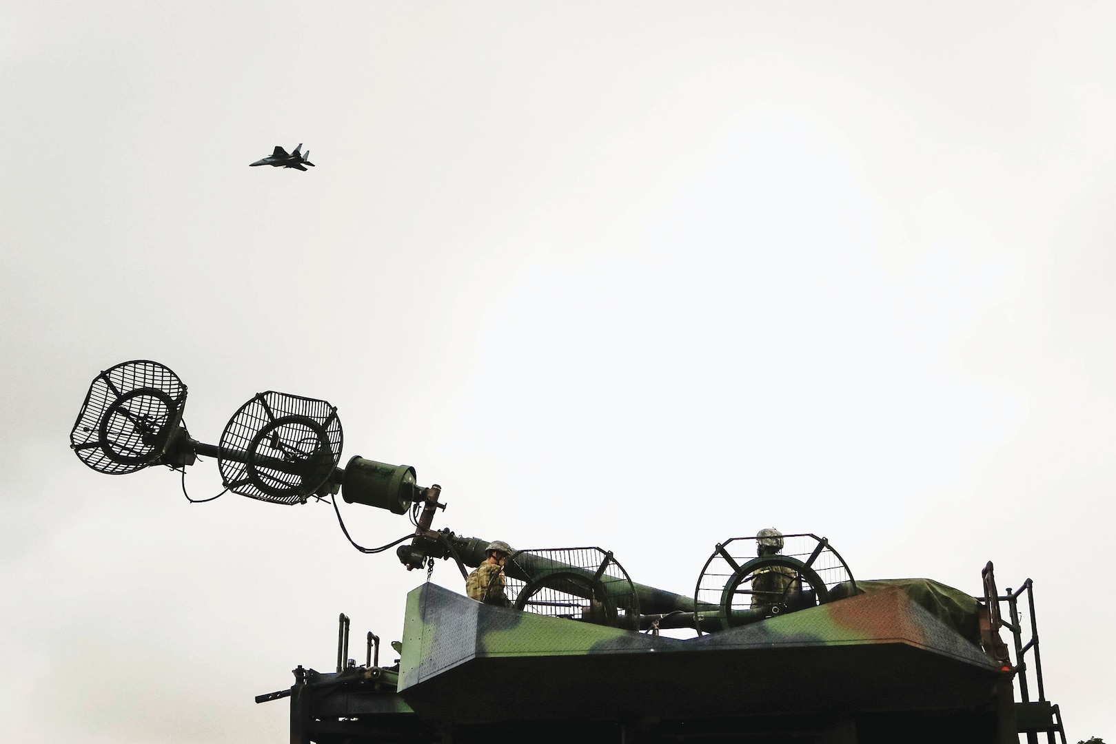 Air Force fighter jet flies overhead as two Soldiers raise signal tower for Patriot missile system