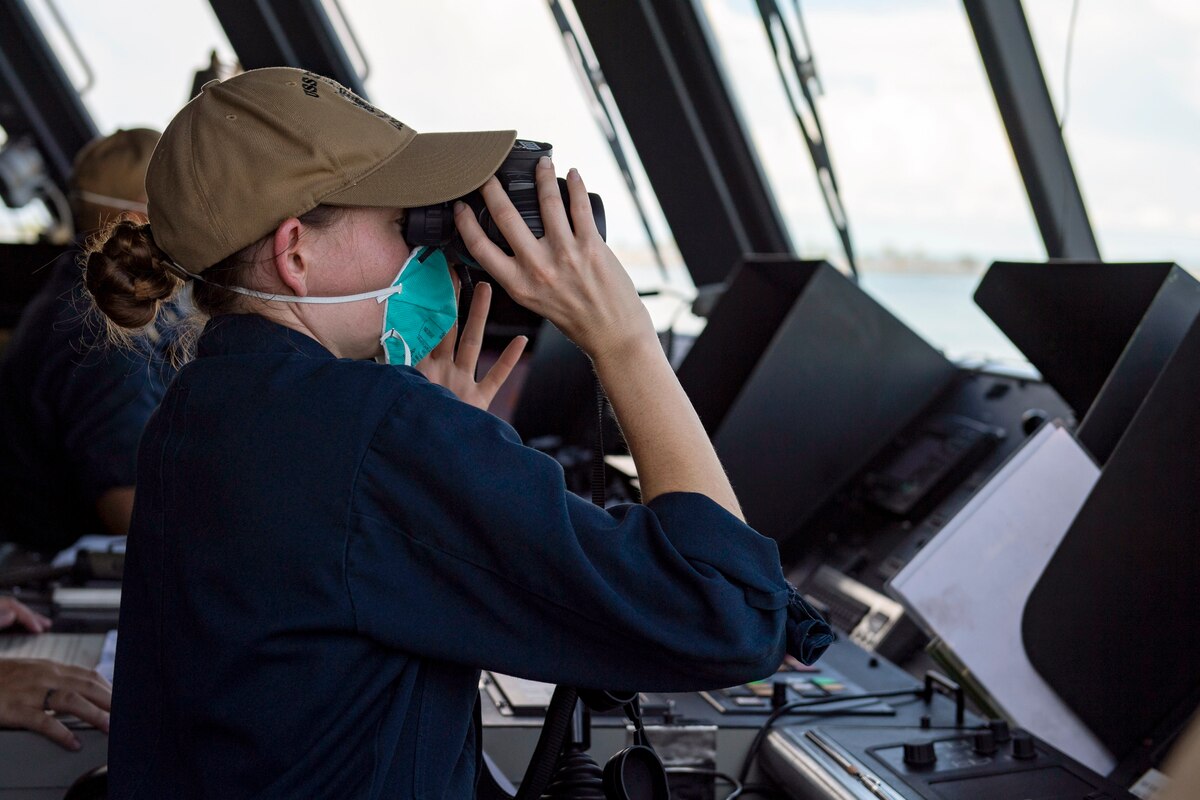 A sailor wearing a mask looks through binoculars while standing in front of a computer screen.
