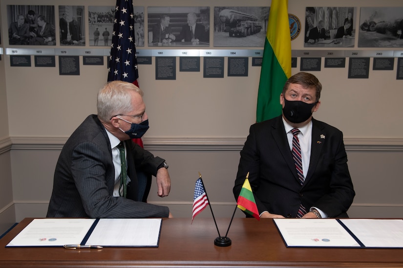 Two men sit next to one another at a table. On the table are pieces of paper, a pen and small Lituanian and U.S. flags.