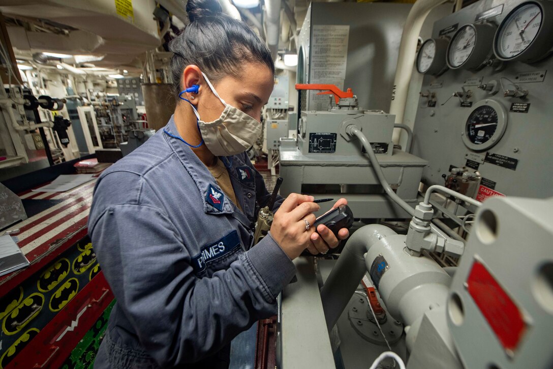 A sailor holds a device while standing the in engine room.