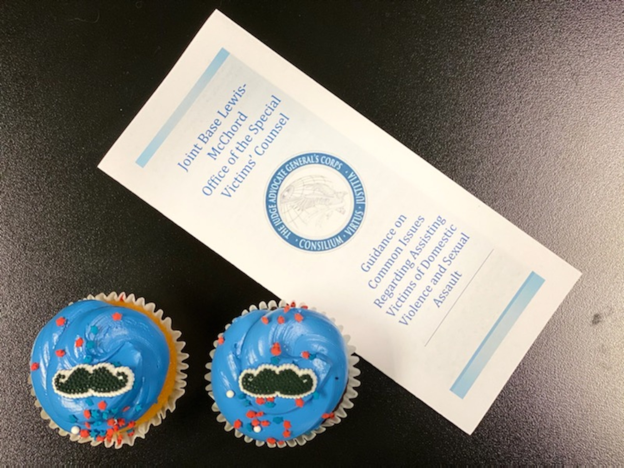 A pamphlet for the Joint Base Lewis-McChord Special Victim's Counsel lies on a table next to blue cupcakes with moustache decorations.