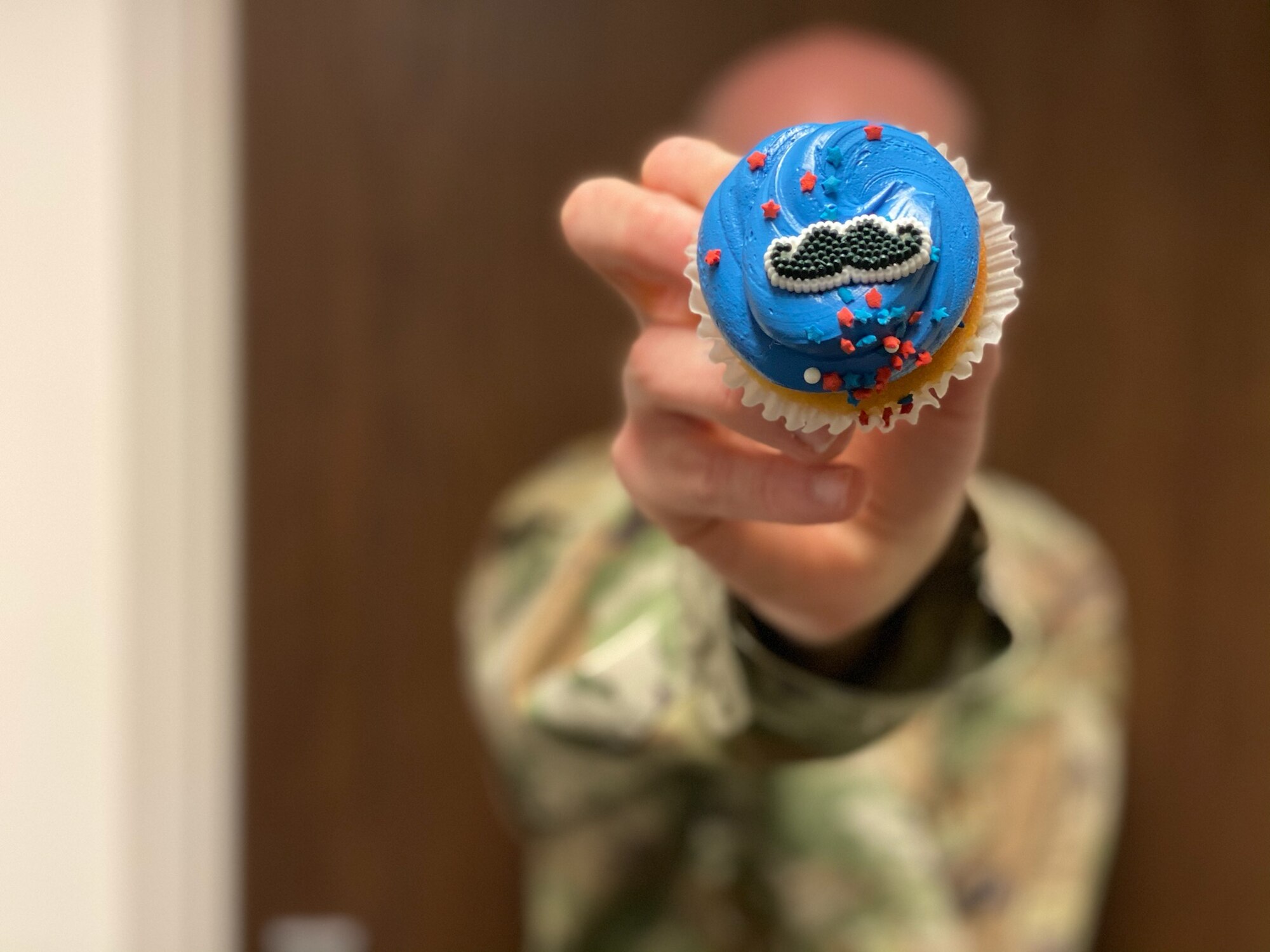 An Airmen holds a cupcake with a moustache decoration up to the camera.