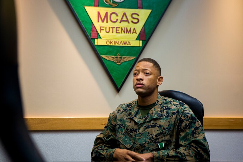 A Marine sits in a chair by a wall hanging of a logo with "MCAS Futenma" written on it.