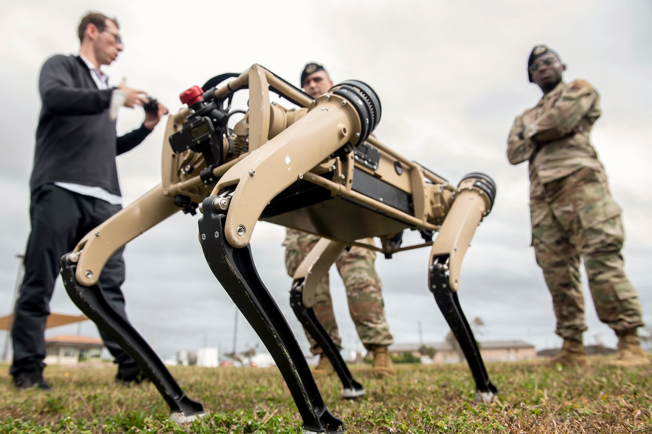 Two airmen stand in a field and look at a four-legged robot as a civilian speaks to them.