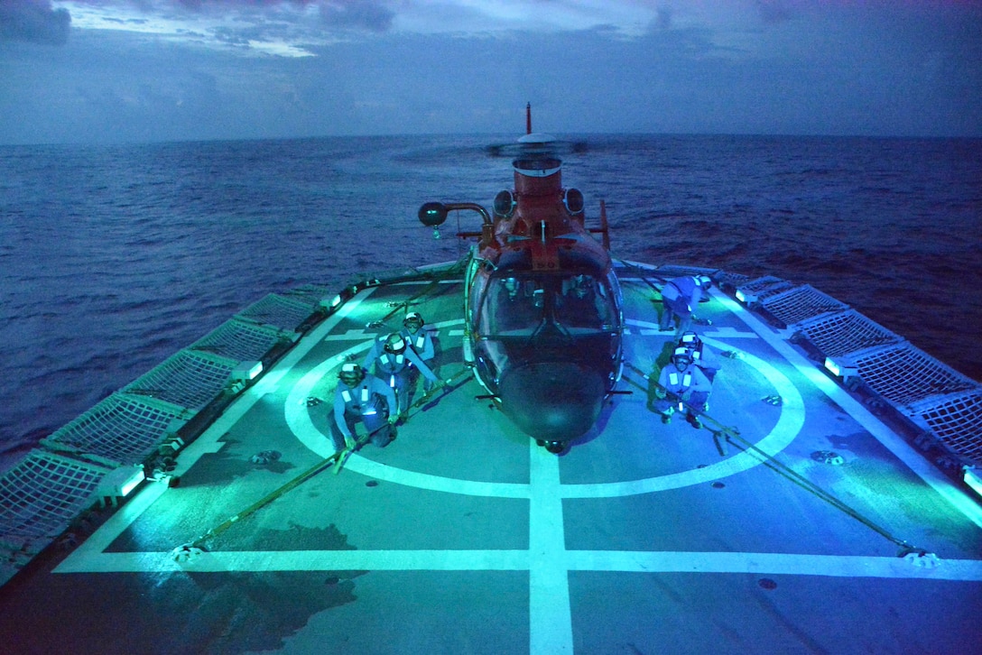 Coast Guardsmen kneel next to a helicopter aboard a ship at sea.