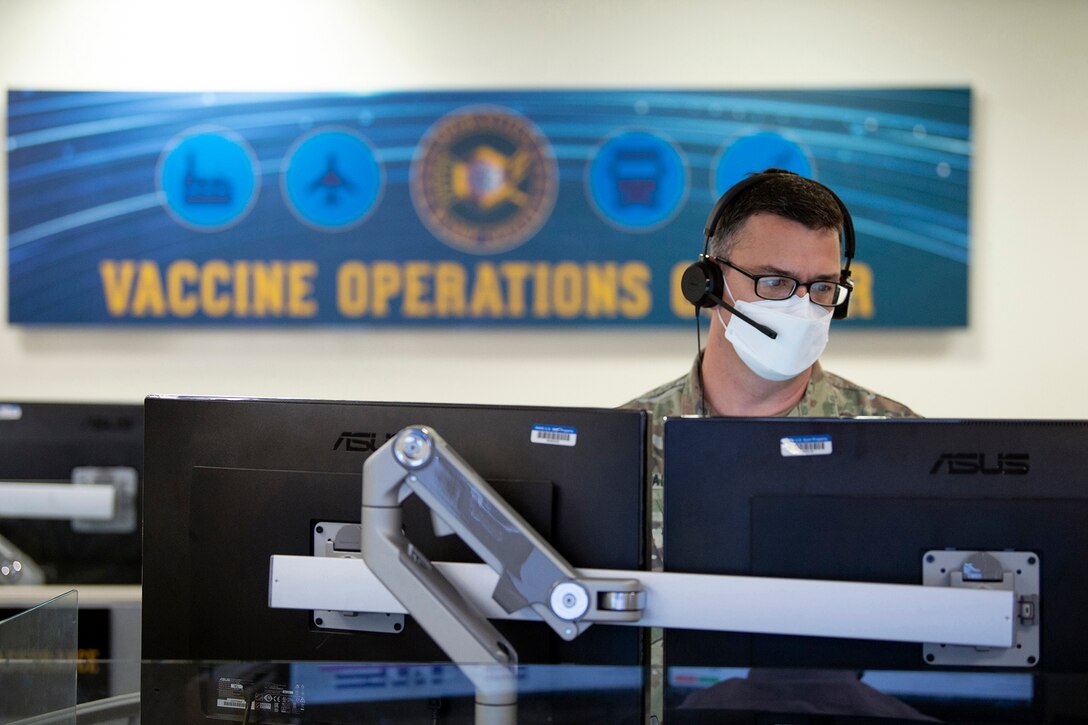 A soldier wearing a face mask works at two computer monitors.