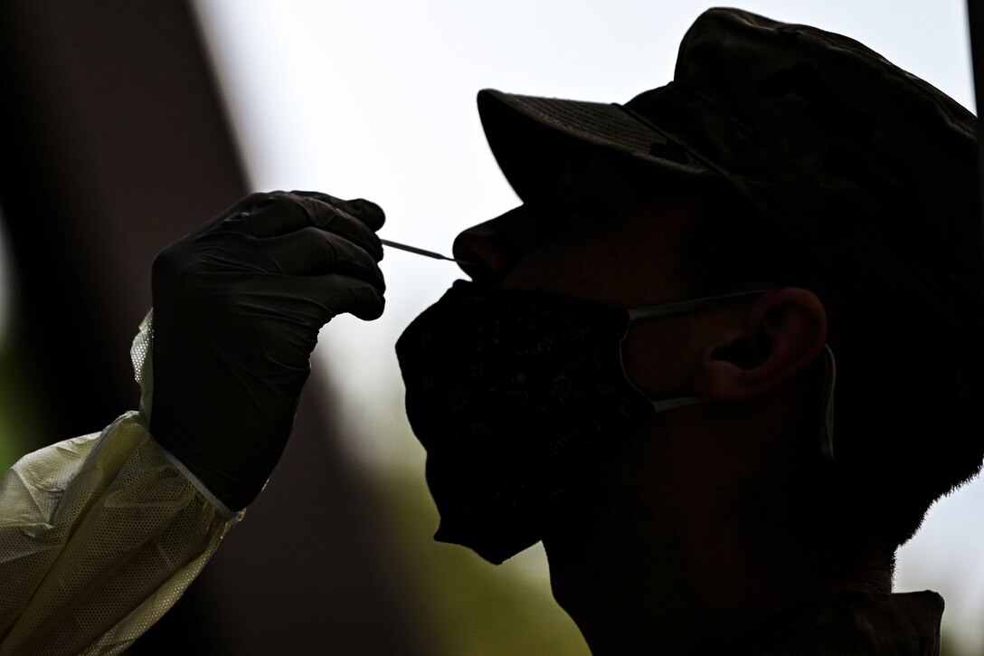 An airman kneels as a service member wearing personal protective equipment swaps his nose.