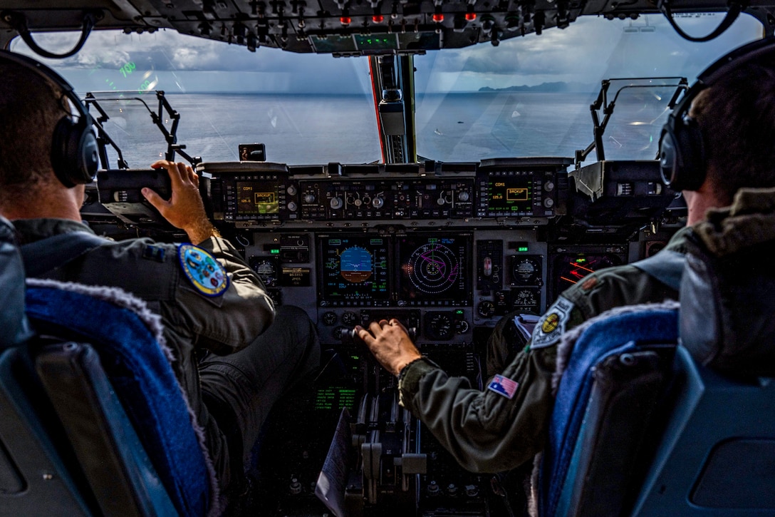 Two pilots, shown from behind, sit in the cockpit of an aircraft flying over water.