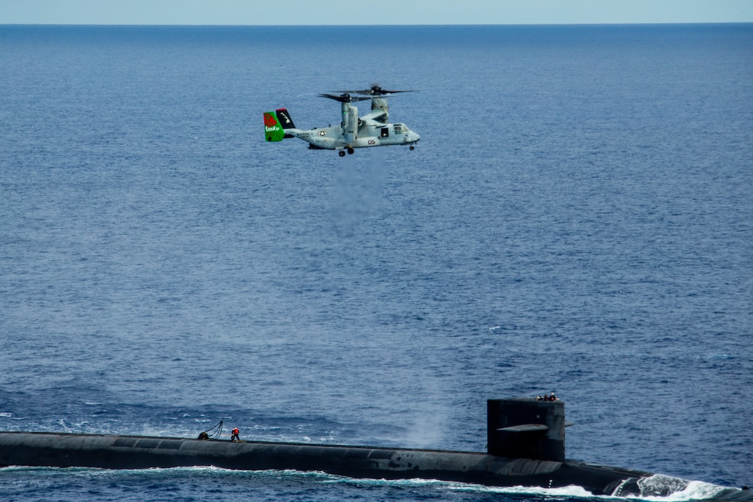 A Marine Corps Osprey aircraft flies above a Navy submarine in blue waters.