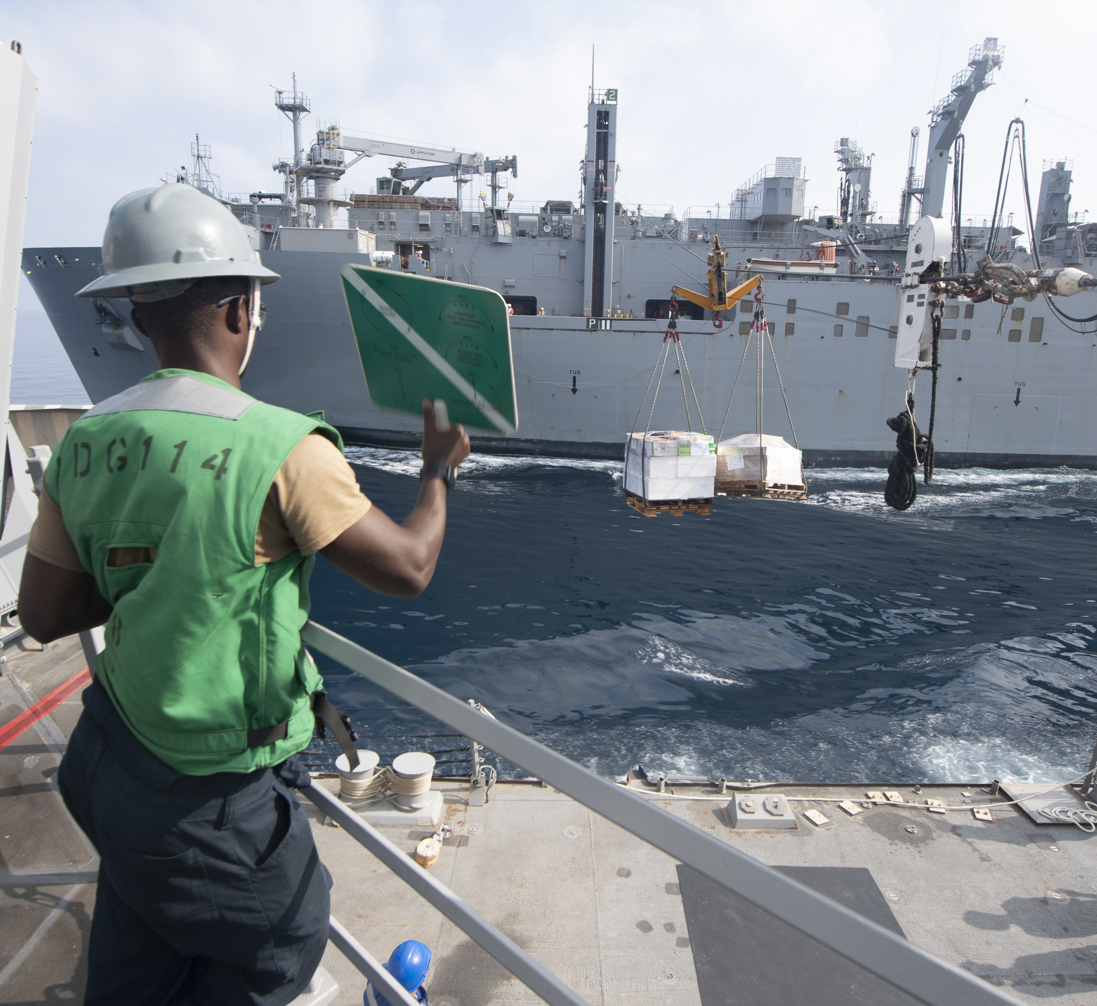 Seaman Allan Wandiri, from Los Angeles, assigned to the guided-missile destroyer USS Ralph Johnson (DDG 114), signals the dry cargo and ammunition ship USNS Wally Schirra (T-AKE 8) during a replenishment-at-sea.