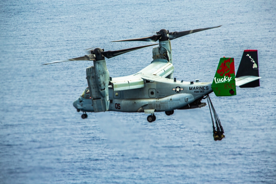 A Marine Corps Osprey aircraft flies with a cargo suspended beneath it.