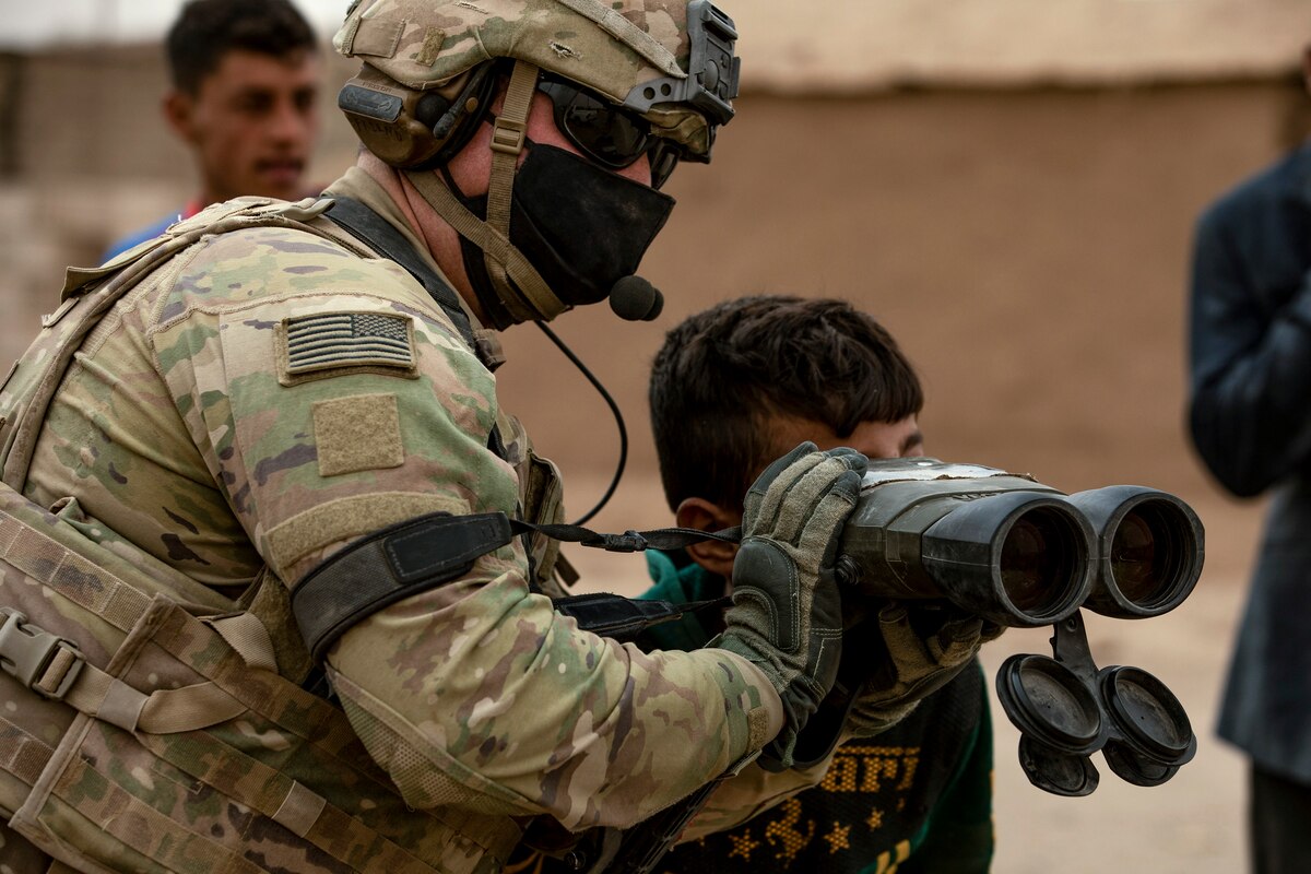 A soldier holds his binoculars as a child looks through them near some buildings outdoors.