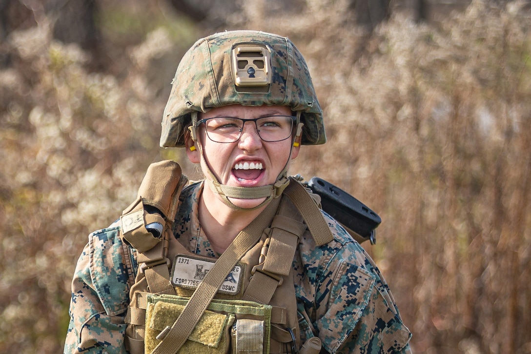 A Marine standing near brush and wearing camouflage and a helmet opens her mouth while shouting.