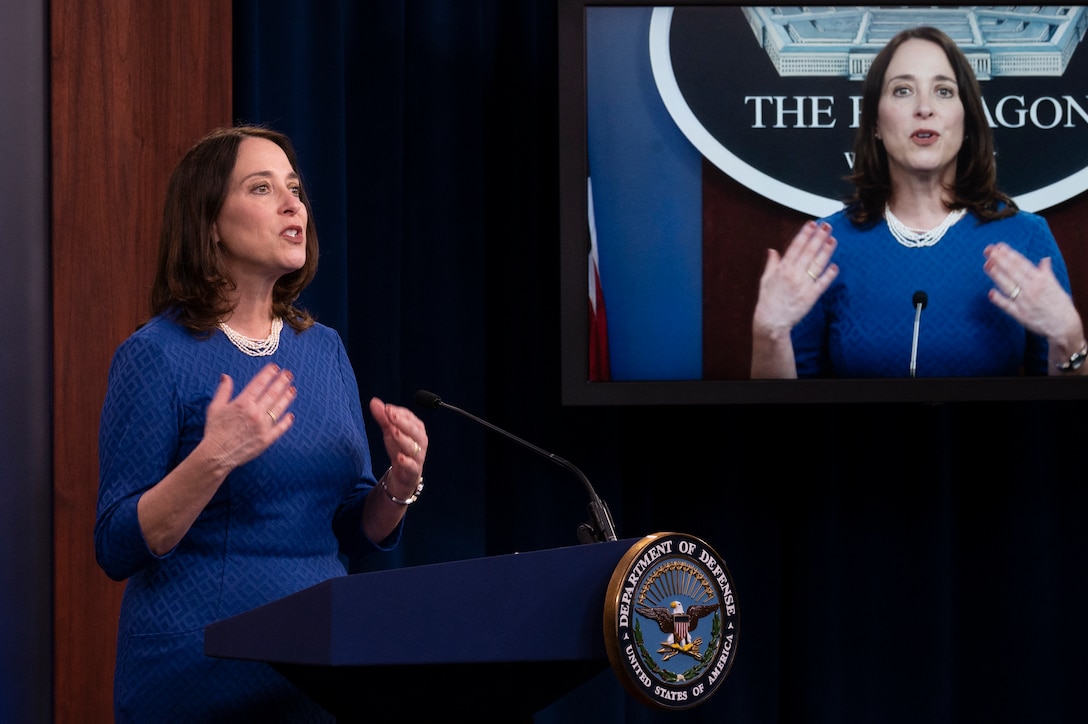 A woman stands behind a lectern. Behind her is a video screen, which also  displays her image.