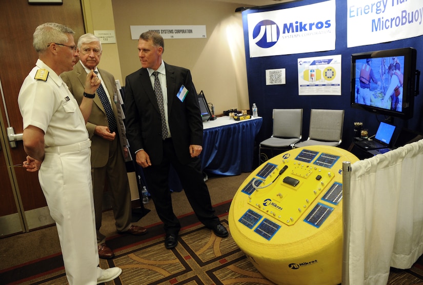 Three men, including one in a military uniform, stand in front of a piece of simulated military hardware at a trade show.